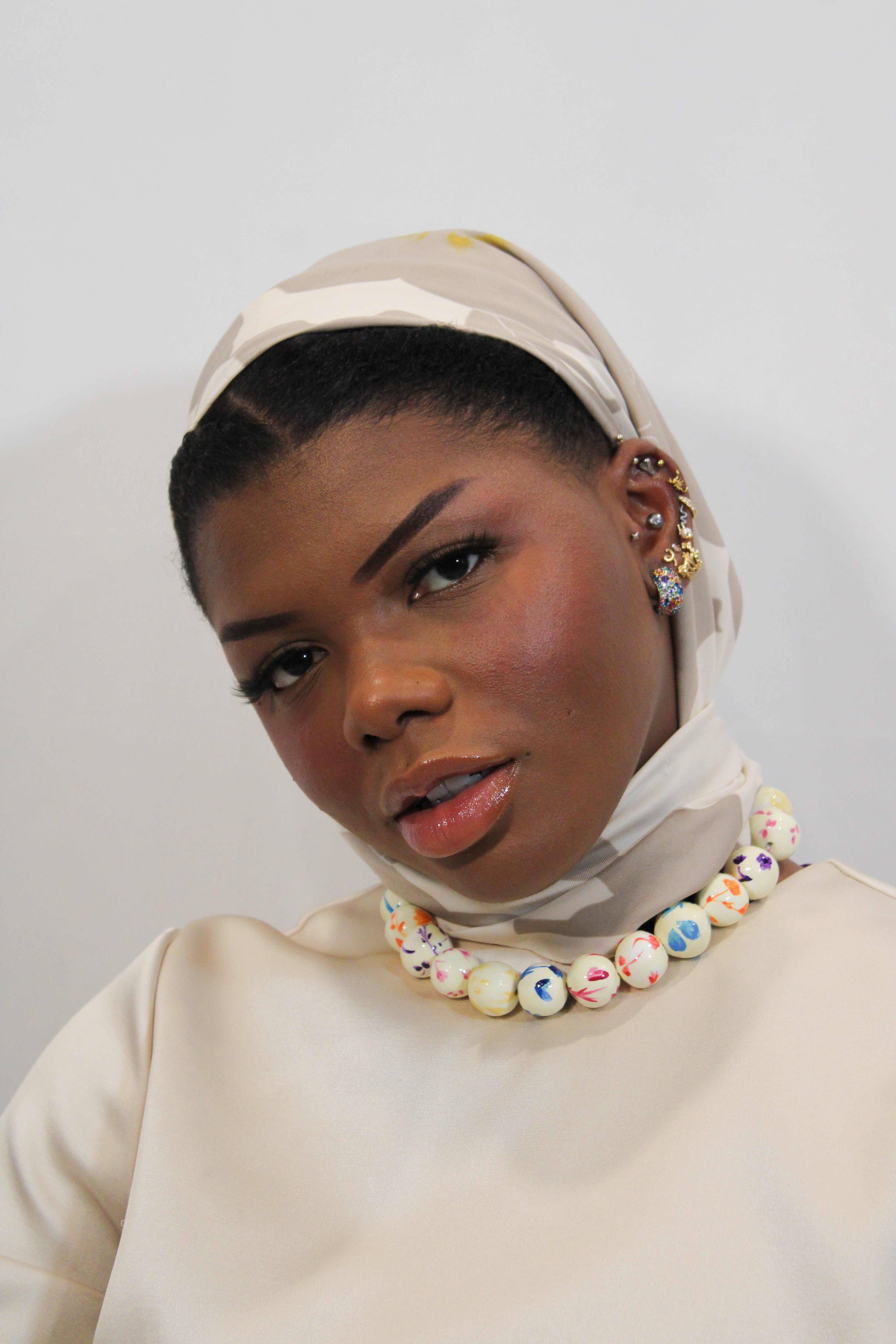 A close-up portrait of a model wearing a cream-colored satin headscarf, a matching cream satin top, and a chunky beaded collar necklace with colorful glass beads.