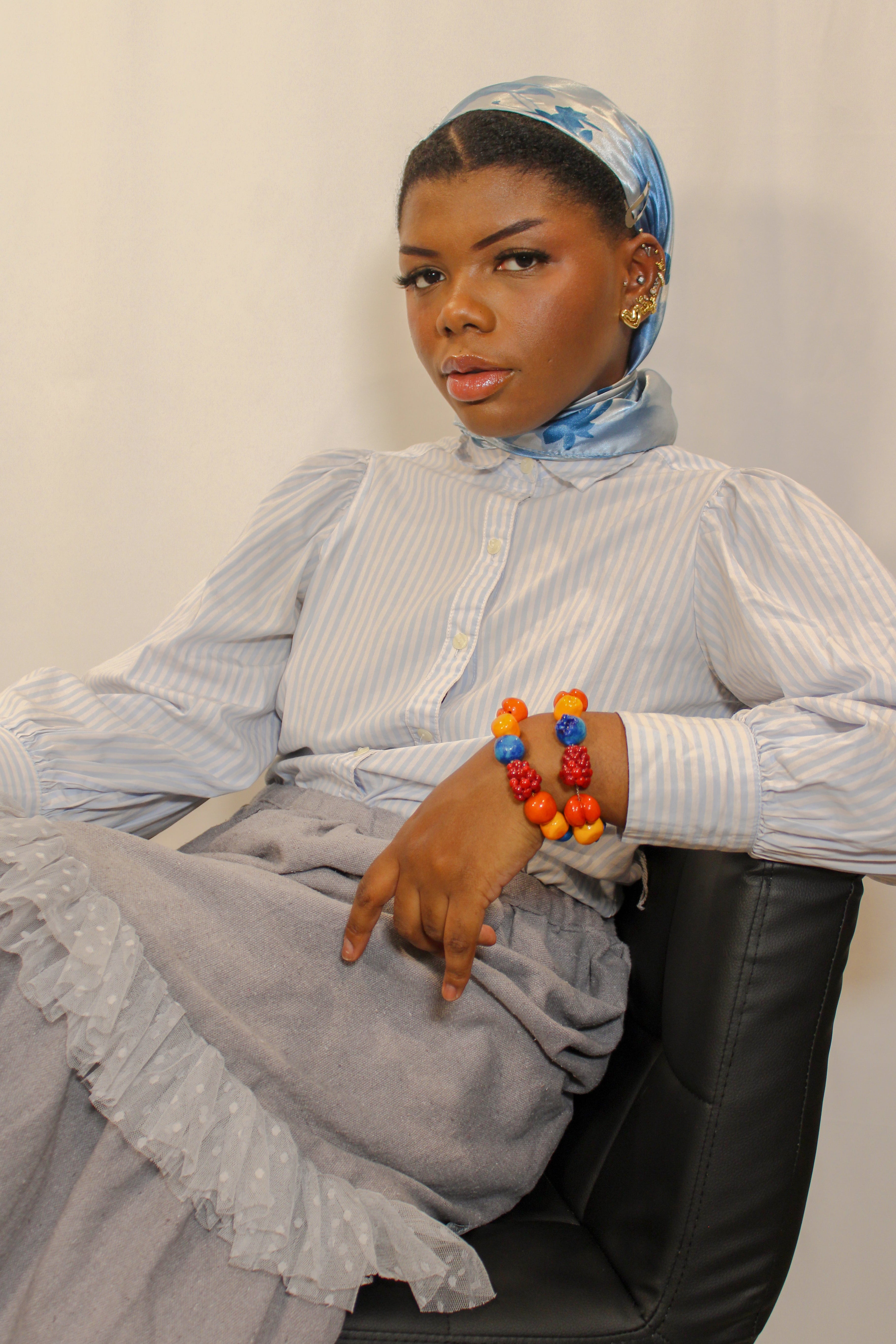 A model wearing a light blue and white striped long-sleeve blouse, a gray ruffled skirt, and a blue patterned headscarf sits in a black chair. She is accessorized with colorful beaded bracelets.